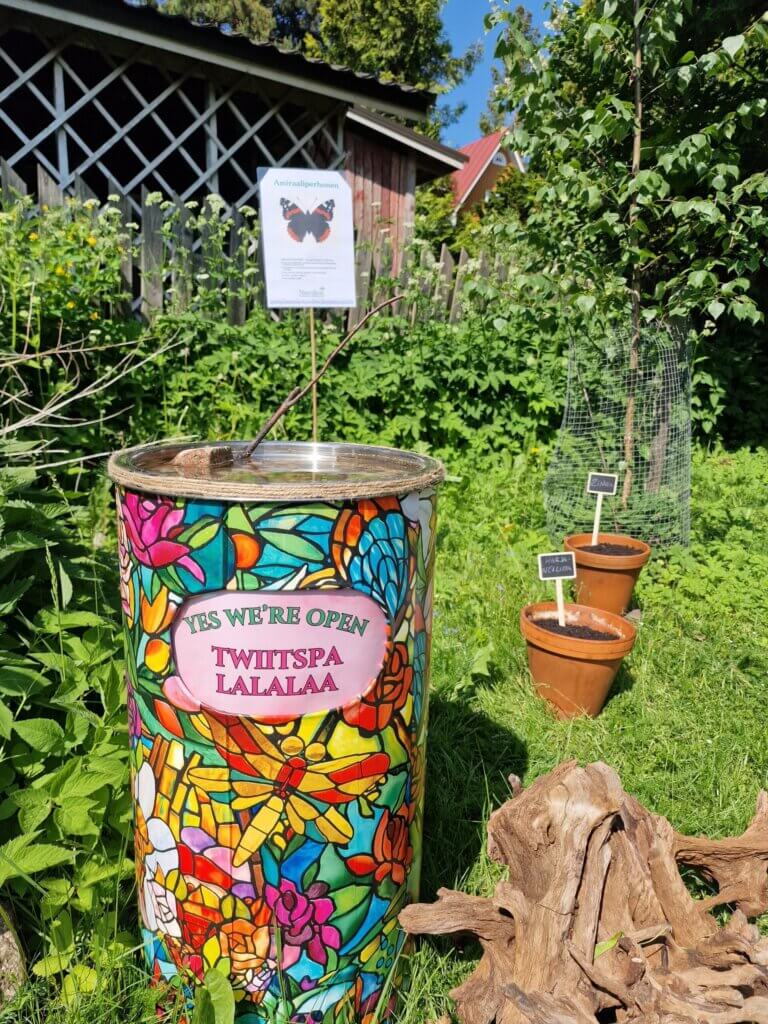 Värikäs kukkakuviollinen astia, jossa on kyltti "Yes we're open, Twiitspa Lalalaa", taustalla puutarha ja infotaulu amiraaliperhosesta.