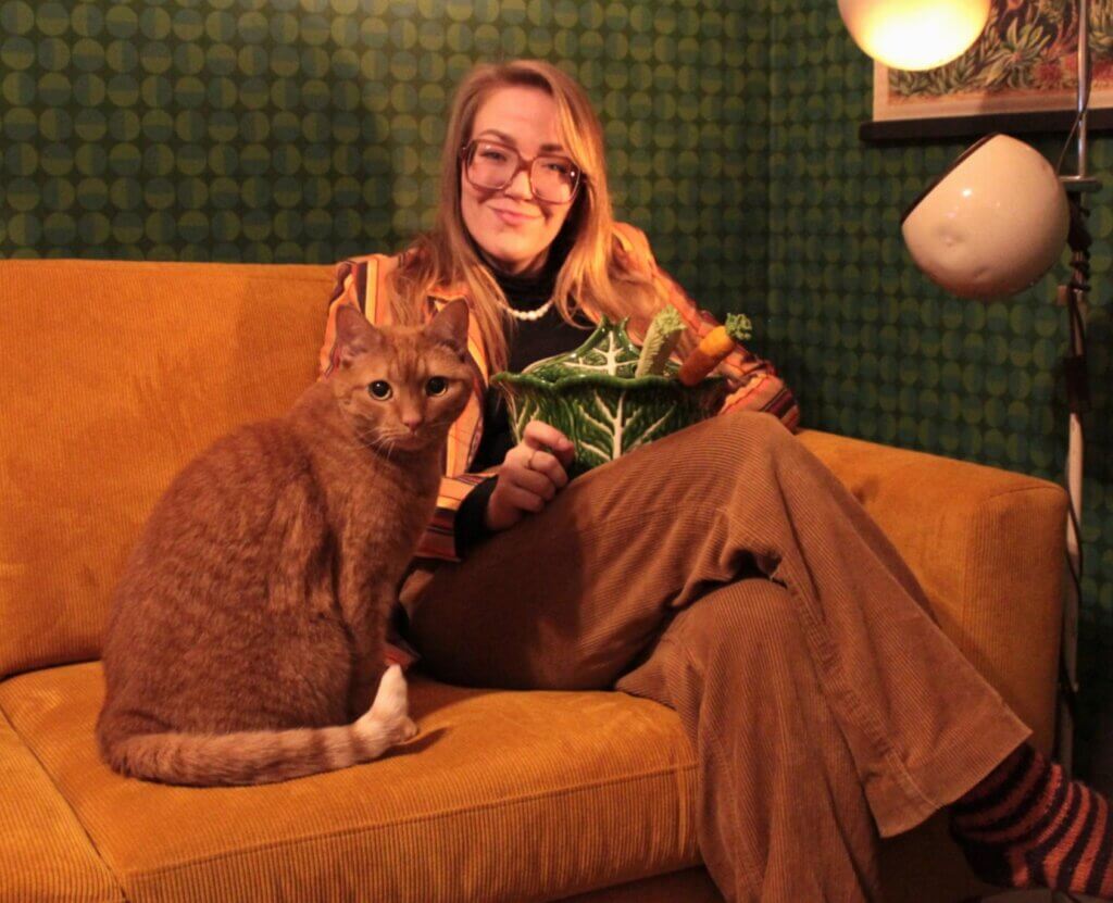 Nainen istuu oranssilla sohvalla, pitelee vihreää kaalin muotoista astiaa ja hänen vieressään on ruskea kissa.