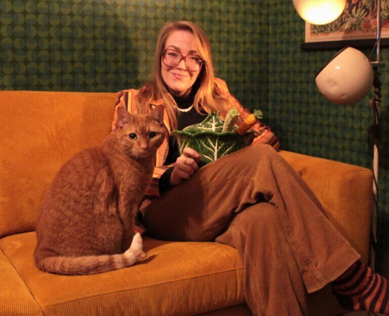 Nainen istuu oranssilla sohvalla, pitelee vihreää kaalin muotoista astiaa ja hänen vieressään on ruskea kissa.