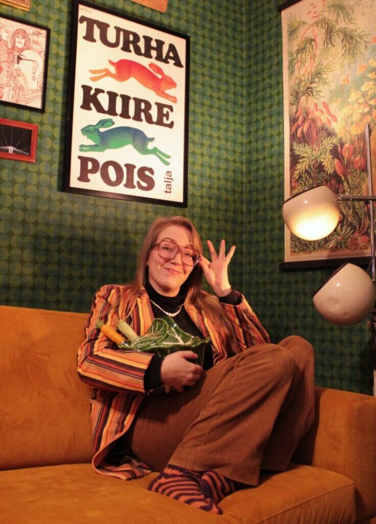 Nainen istuu oranssilla sohvalla, pitää käsissään vihanneksia ja hymyilee kameralle, taustalla on värikäs juliste ja koristeellista tapettia.