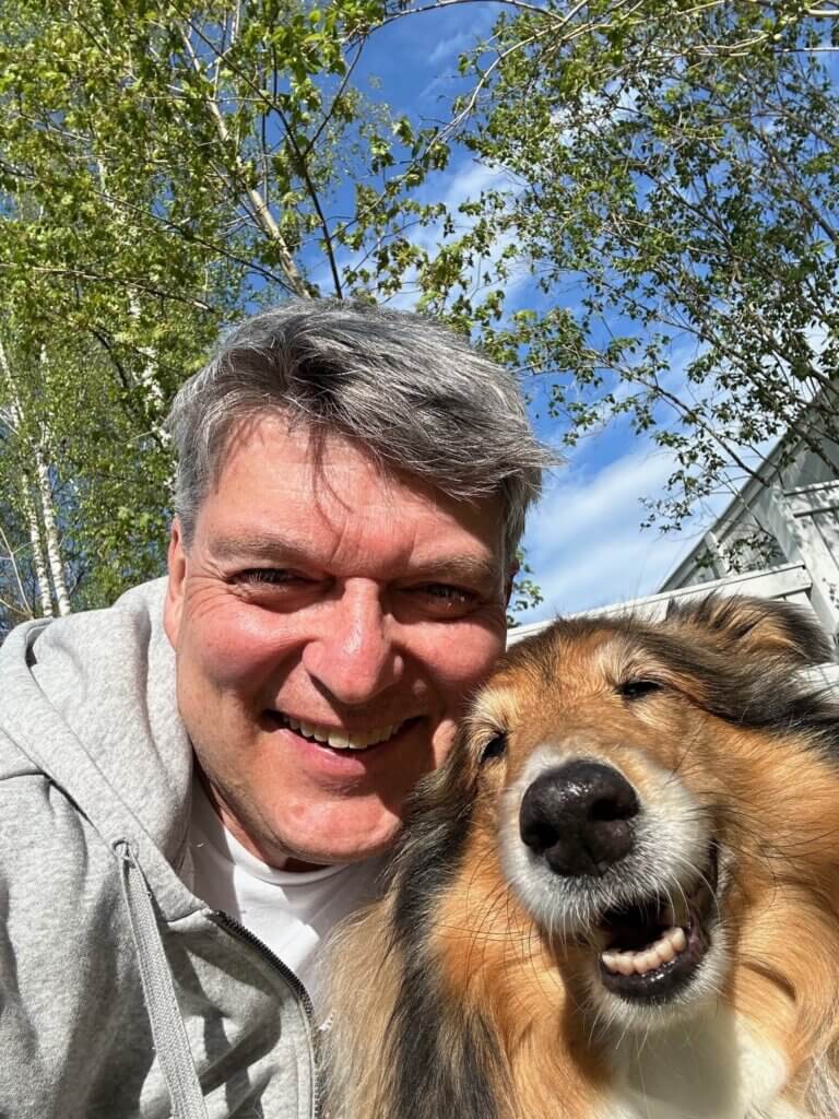 Mies hymyilee ottaessaan selfie-kuvaa koiransa kanssa ulkona puiden taustalla.