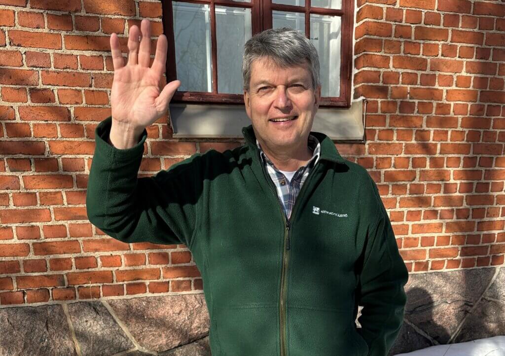 Mies hymyilee ja heiluttaa kättään punatiiliseinän edessä.