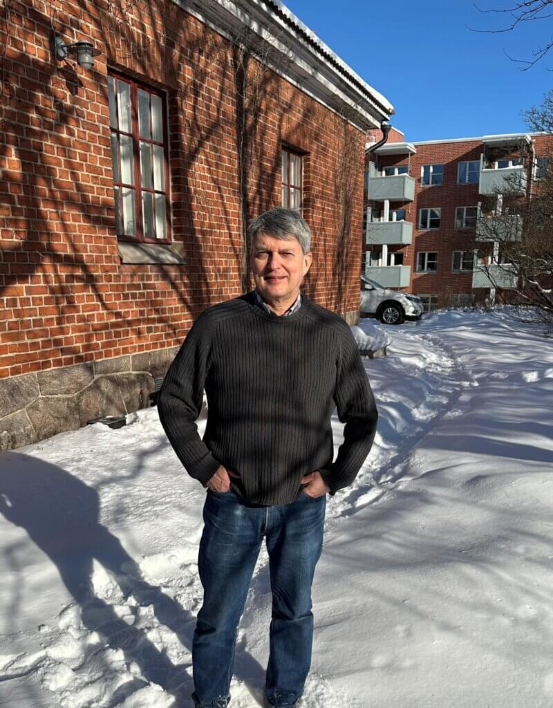 Mies seisoo lumisessa maisemassa tiiliseinän edessä auringonpaisteessa.