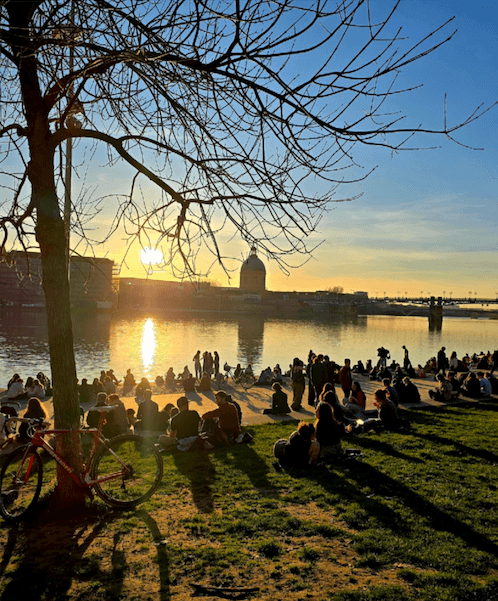 Auringonlasku heijastuu vedessä, kun ihmiset nauttivat kevätsateista puistossa puun alla.
