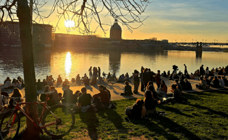 Aurinkoinen ilta-aurinko heijastuu joen pinnasta, kun ihmiset nauttivat ulkona istumisesta ja seurustelusta.