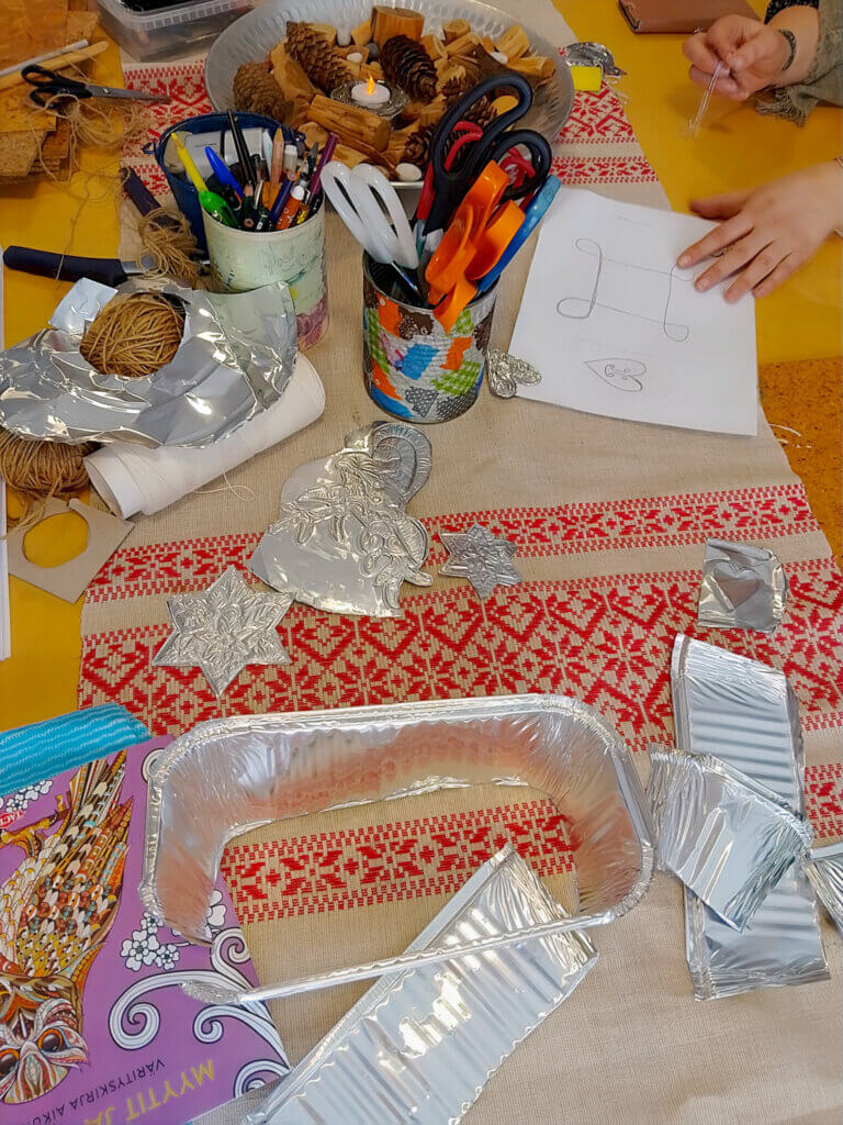 Työpöydällä on erilaisia askartelutarvikkeita, kuten kyniä, sakset, alumiinifoliota ja paperia, sekä koristeita ja luonnonmateriaaleja.