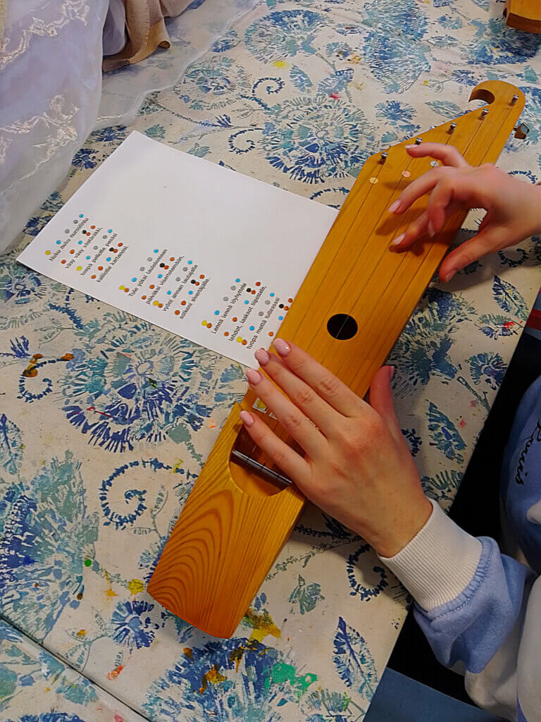 Kuvassa on henkilö, joka soittaa puista kanteletta ja katsoo nuottilehteä, jossa on värikoodatut merkinnät.