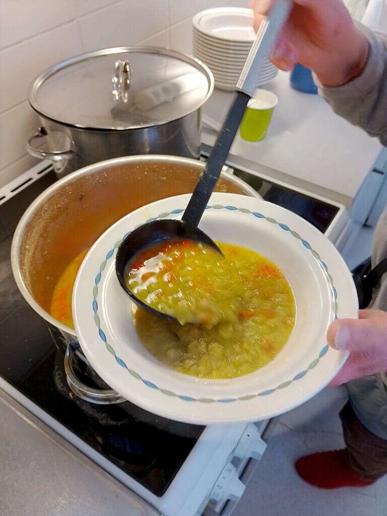 Käsi kaataa keittoa lautaselle mustalla kauhalla kattilasta, taustalla näkyy astioita.