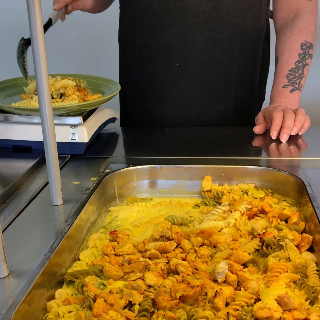 Kuvassa on henkilö, joka annostelee pastaa ja kanaa lautaselle keittiössä.