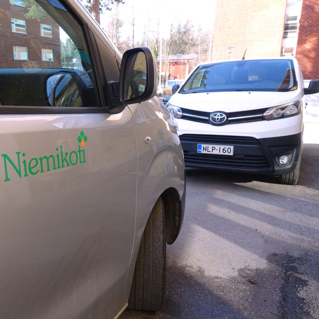 Kuvassa on kaksi autoa, joista toinen on harmaa ja siinä on "Niemikoti"-logo, ja toinen on valkoinen Toyota.