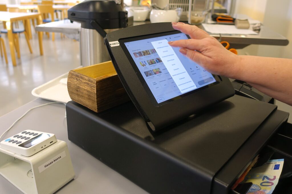 Kuvassa näkyy kassa, jossa henkilö käyttää kosketusnäyttöä ja taustalla on rahaa sekä kahvinkeitin.