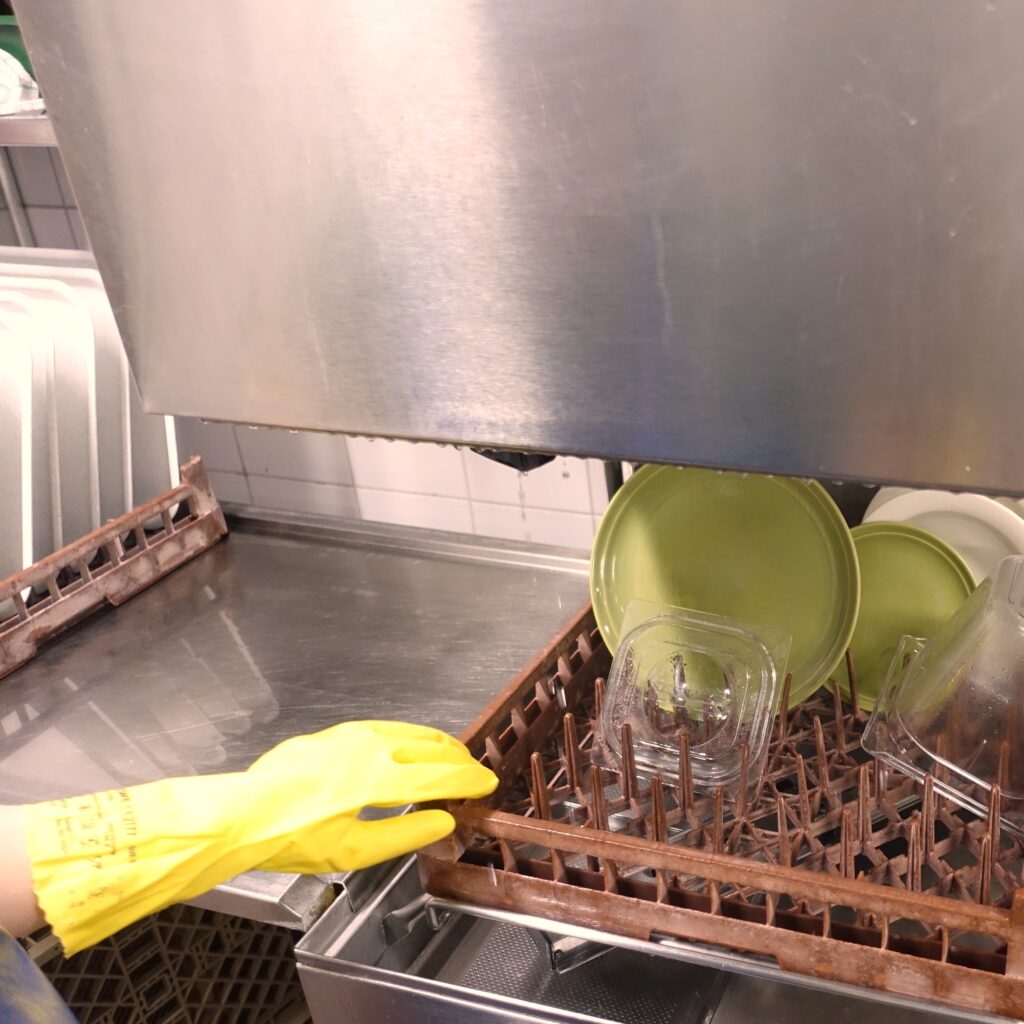 Kuvassa näkyy käsin peseminen astioita teollisessa astianpesukoneessa, jossa on keltainen kumihansikas.