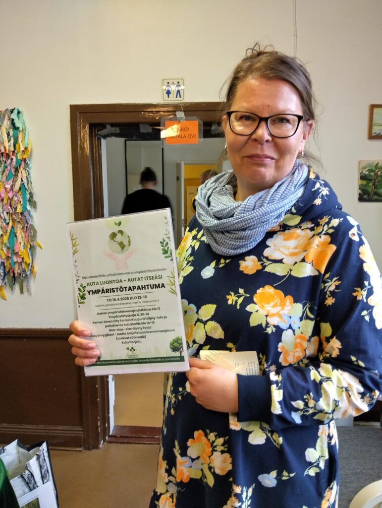Nainen pitää kädessään esitteitä ympäristötapahtumasta ja hymyilee kameralle.