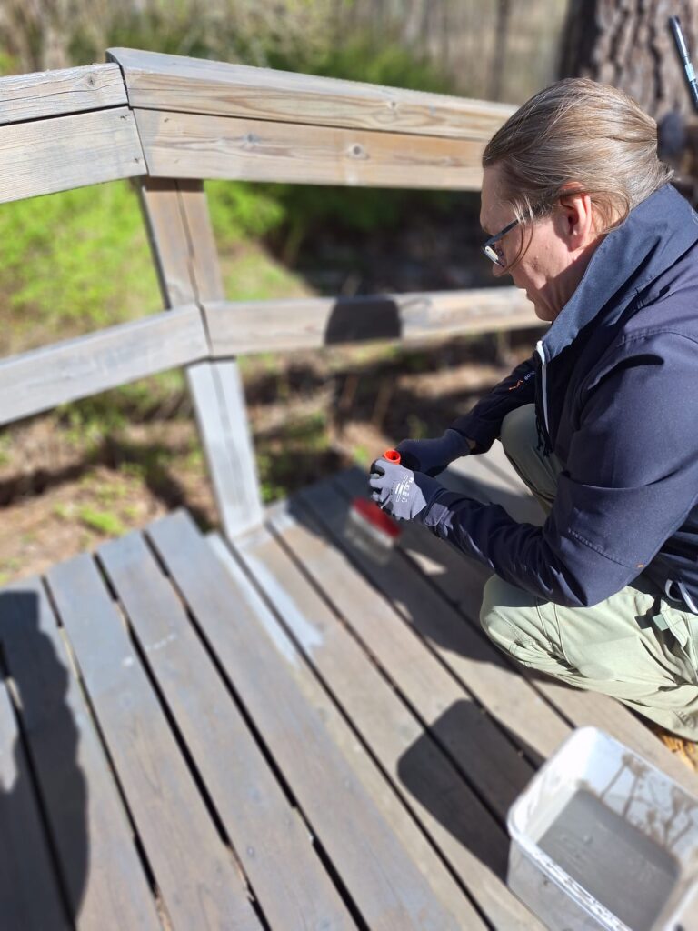 Henkilö maalaa puista terassia ulkona, käyttäen harjaa ja suojakäsineitä.