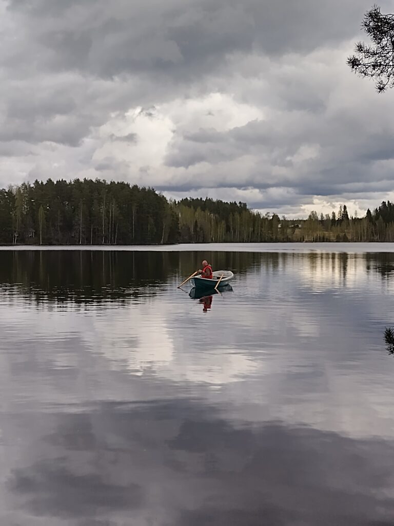 Mies soutaa veneellä tyynellä järvellä, taustalla tummia pilviä ja metsää.