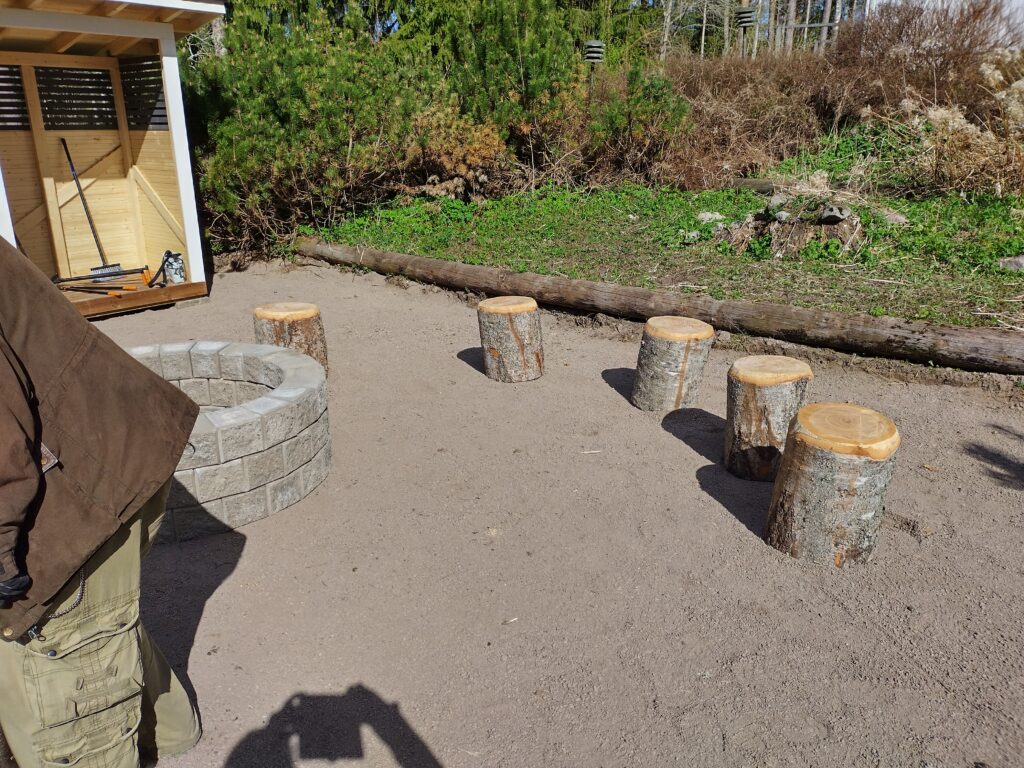 Kuvassa on nuotiopaikka kivikehällä ja puupölkkyjä istuimina metsäisessä ympäristössä.