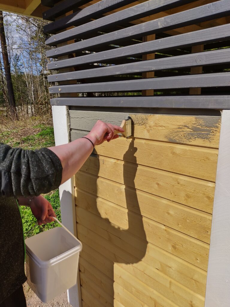 Henkilö maalaa puuseinää pienellä telalla ja pitää toisessa kädessä valkoista maalausämpäriä.