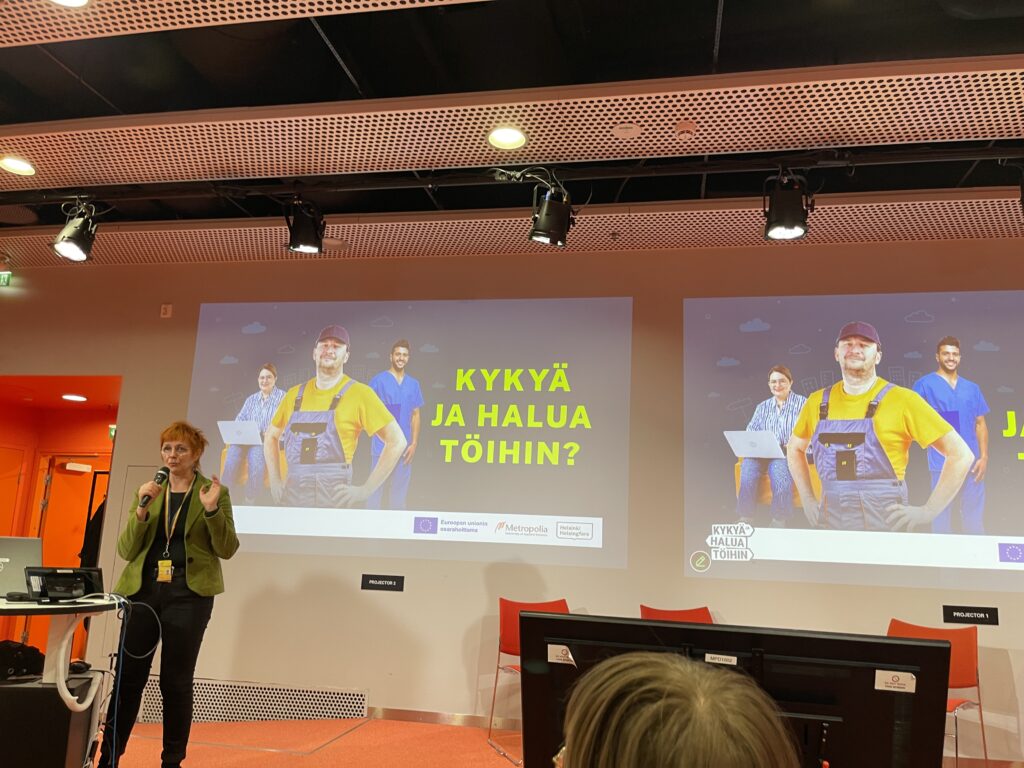 Esitystilanteessa nainen puhuu yleisölle, taustalla on kolme henkilöä eri ammateissa ja suuri kysymys "KYKYÄ JA HALUA TÖIHIN?" näyttötaululla.