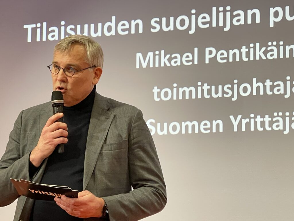 Mikael Pentikäinen puhuu mikrofonin kautta tapahtumassa, taustalla näkyy teksti "Tilaisuuden suojelijan puhe".