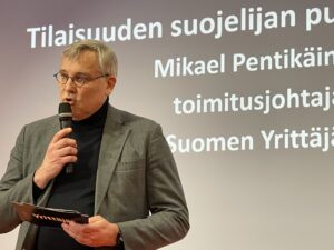 Mikael Pentikäinen puhuu mikrofonin kautta tapahtumassa, taustalla näkyy teksti "Tilaisuuden suojelijan puhe".