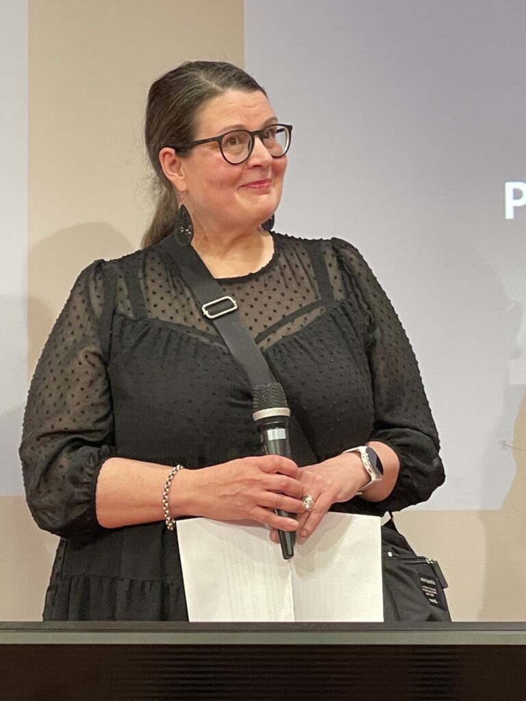 Nainen mustassa, läpikuultavassa paidassa, joka pitää mikrofonia ja katsoo hymyillen yleisöön.