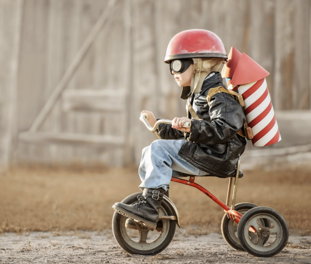Ett barn i en röd hjälm och solglasögon cyklar på en liten cykel med en pappersraket på ryggen.