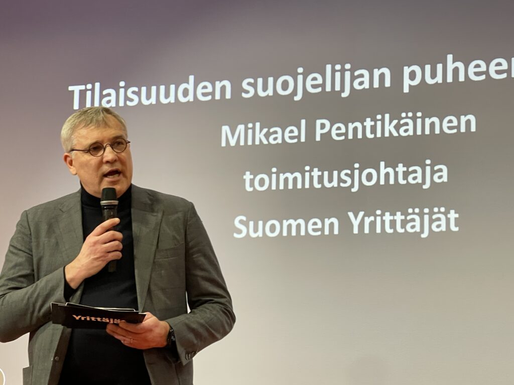 Mikael Pentikäinen puhuu tilaisuuden suojelijana mikrofonin kanssa, taustalla näkyy hänen nimensä ja titteli.