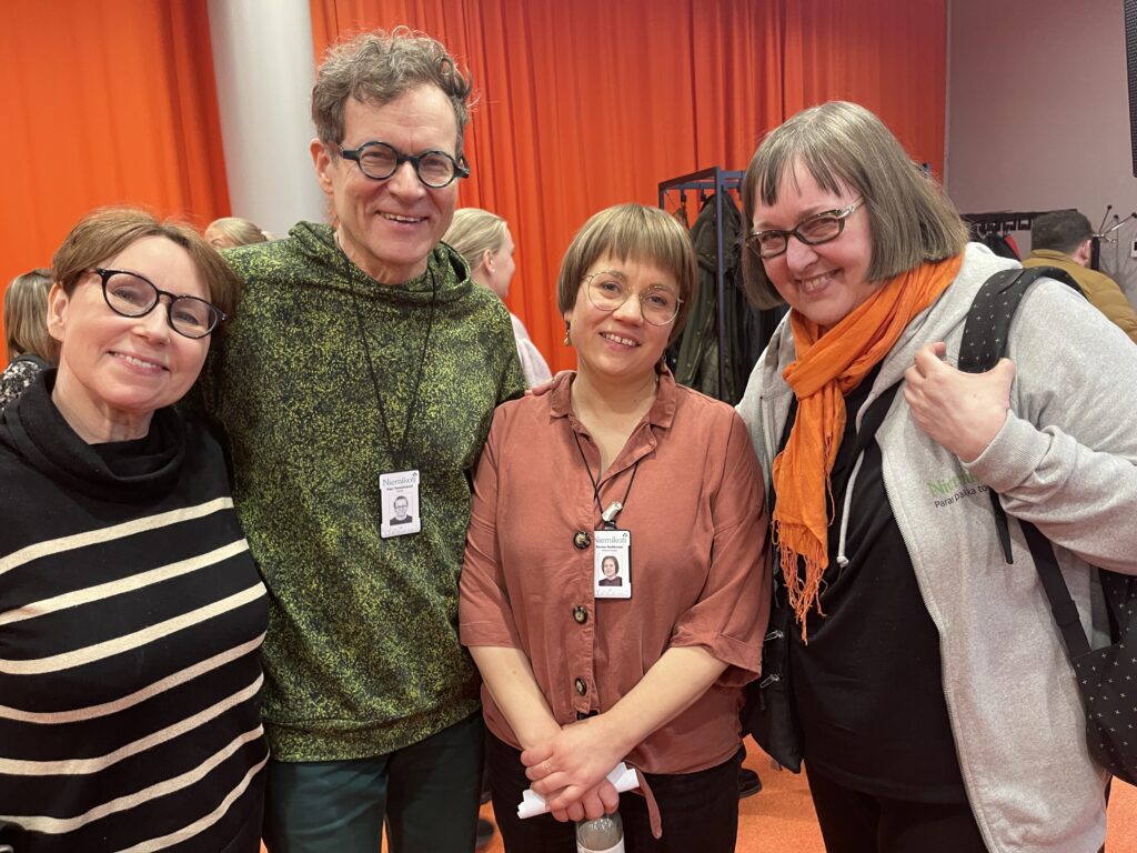 Neljä henkilöä seisoo yhdessä oranssia taustaa vasten, ja heillä on ystävälliset ilmeet.