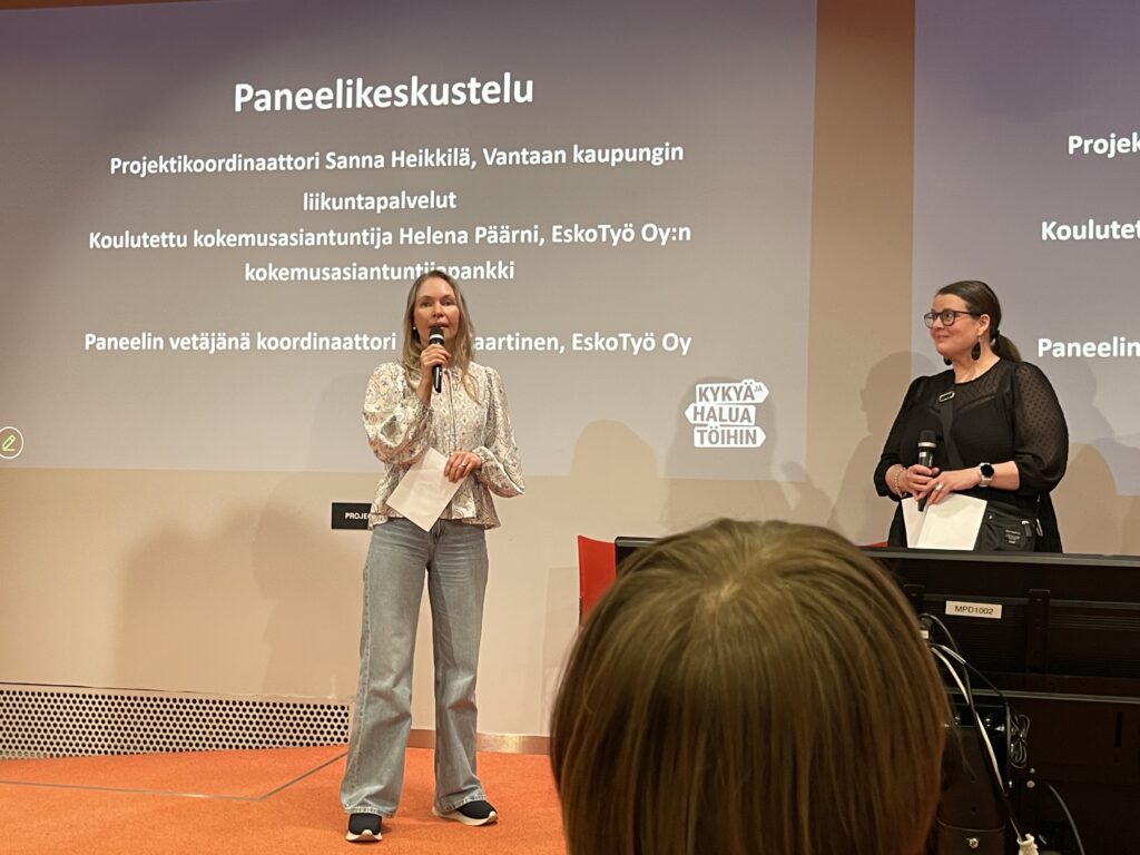 Kuvassa kaksi naista puhumassa paneelikeskustelussa, taustalla esityskalvoja ja mikrofoneja.