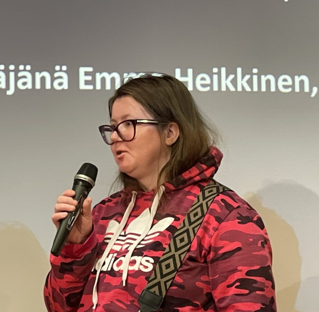 Naispuolinen henkilö puhuu mikrofonin kanssa, taustalla näkyy tekstiä ja toinen henkilö kuuntelee.