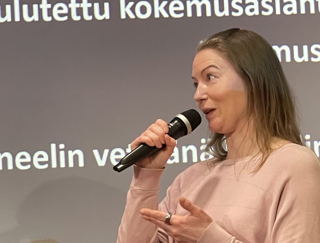 Nainen puhuu mikrofonin kanssa esitystilanteessa, taustalla näkyy tekstiä.