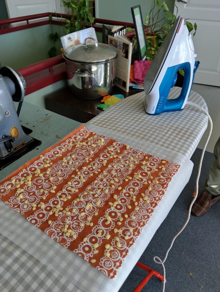 Kuvassa on silitysrauta, joka on asetettu pöydälle, ja sen vieressä on oranssi kangaspala, jossa on vahakappaleita valmistettavaksi.