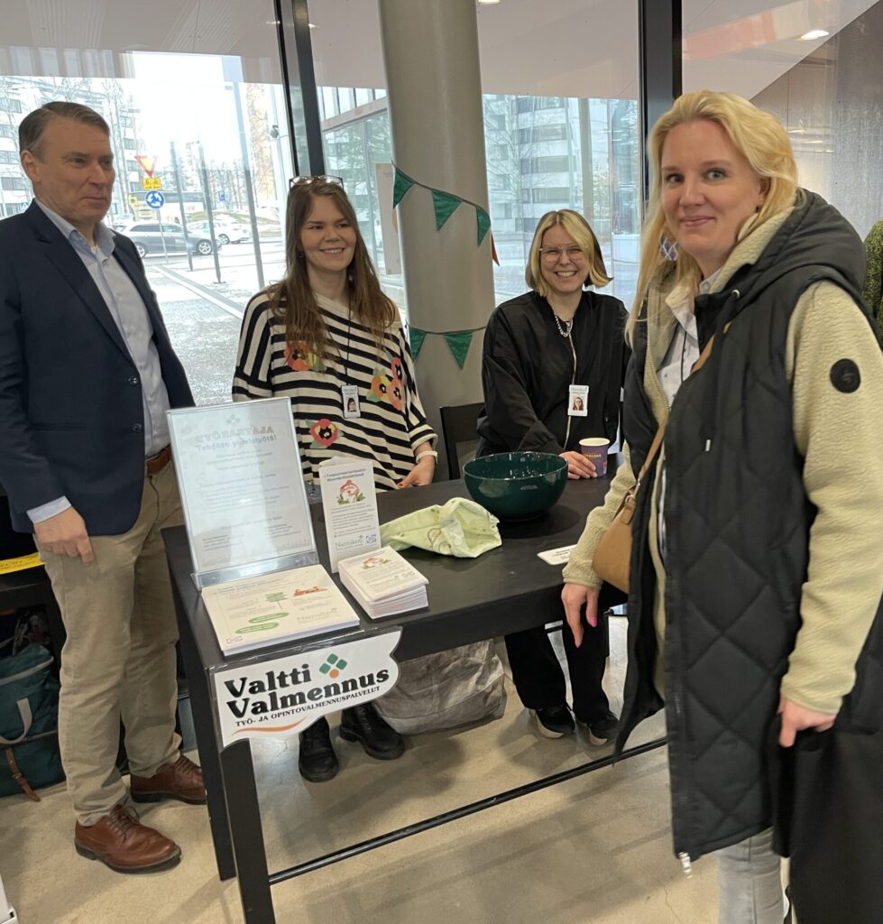 Neljä henkilöä seisoo pöydän äärellä, jossa on esitteitä ja kulho.
