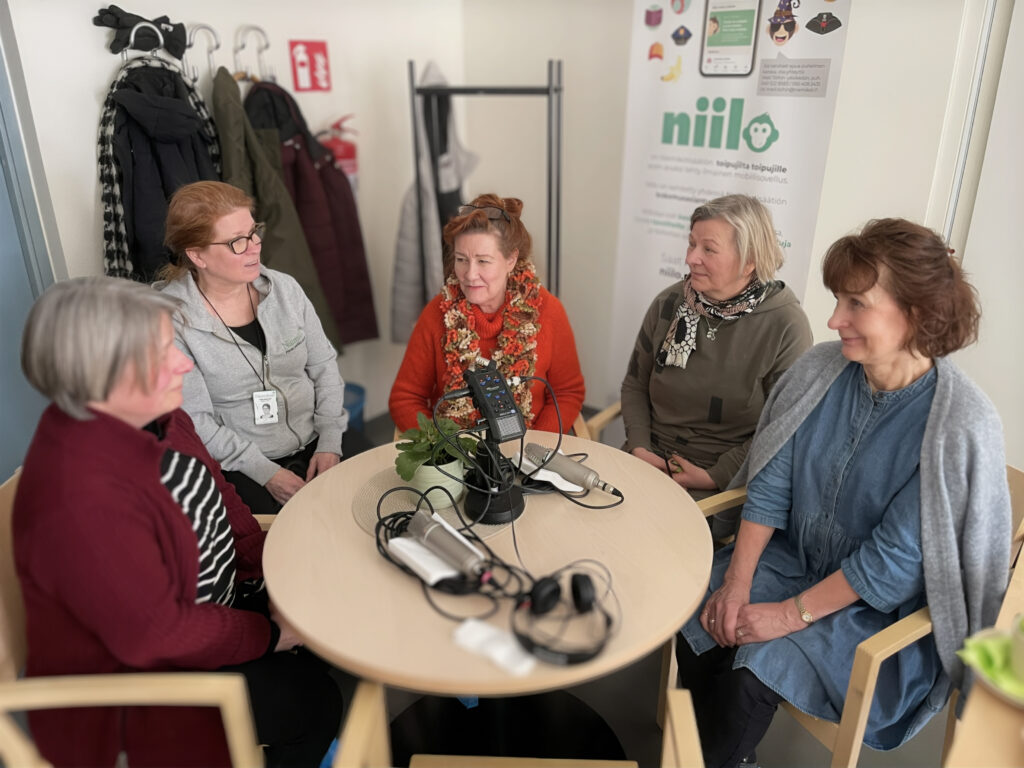 Viisi naista istuu ympyrässä pöydän ääressä keskustelemassa, taustalla näkyy mainos ja vaatteita naulakossa.