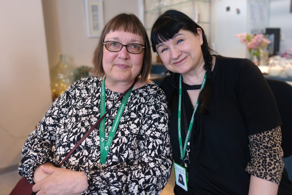 Kaksi naista hymyilee kameralle, päällä kukkakuosiset paidat ja kaulassa nimilaput.