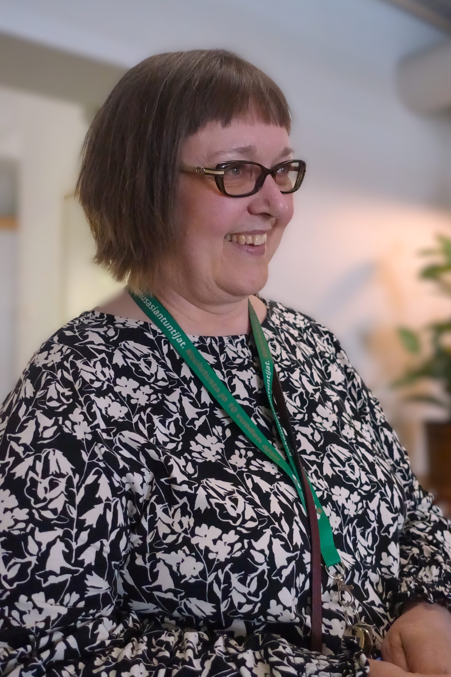 Nainen, jolla on lyhyet hiukset ja silmälasit, hymyilee musta-valkoisessa kukkakuosisessa paidassa ja vihreässä nimikortissa.