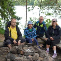 Neljä ihmistä kesäisessä luonnossa nuotiopaikan ympärillä taustalla järvi